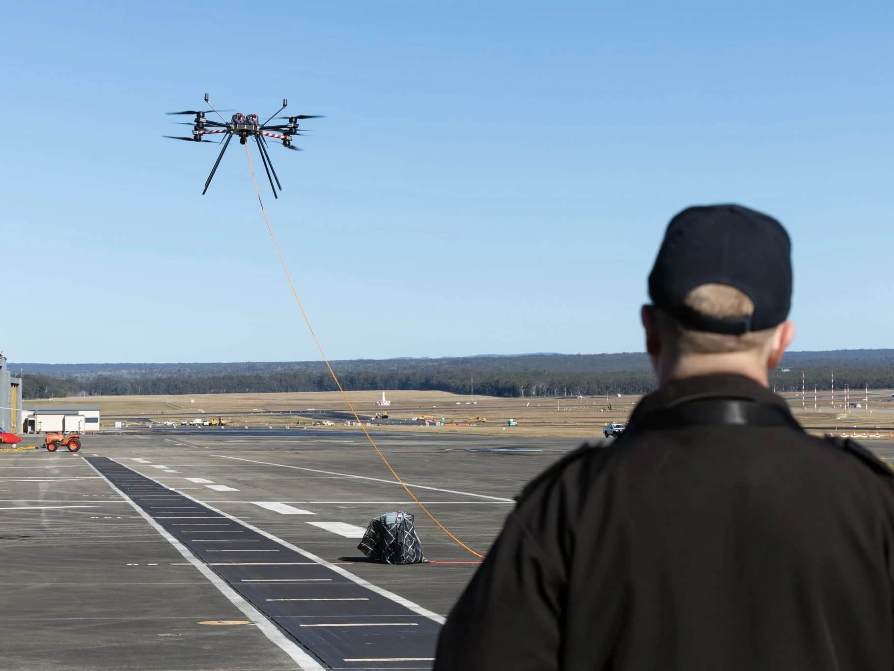 A Callisto in flight over an airfield apron, a line running from it to a load on the ground, watched by a Royal Australian Navy officer in the foreground