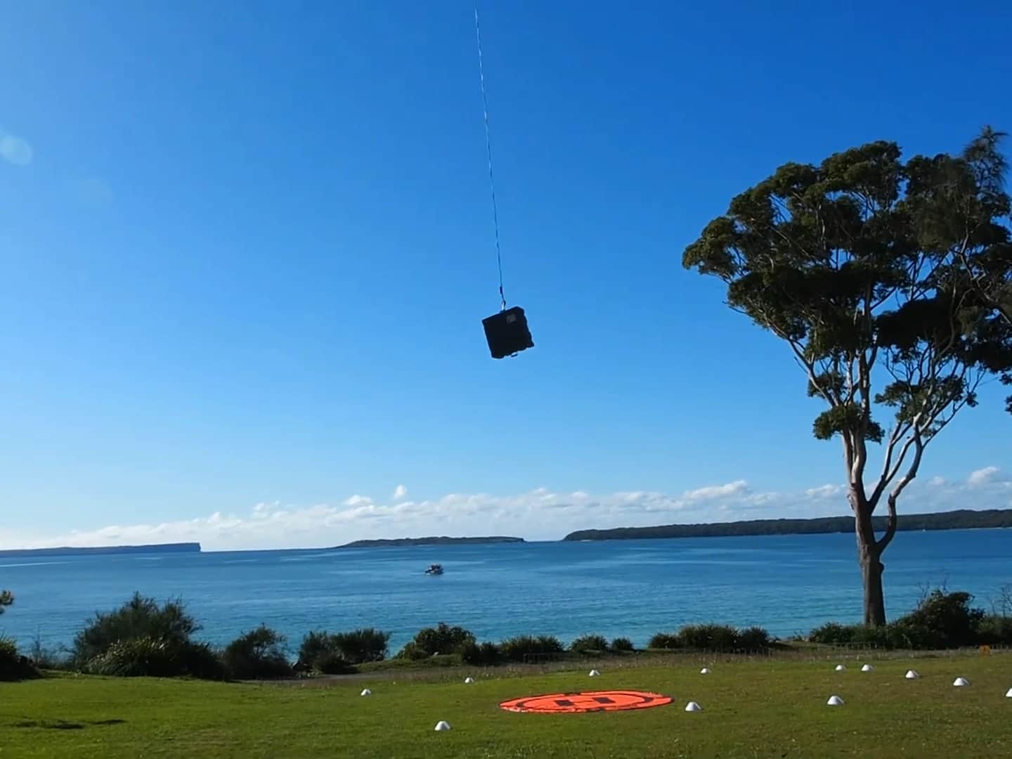 A Callisto climbing away from its landing pad above a bay with a load beneath it, a vessel out on the water